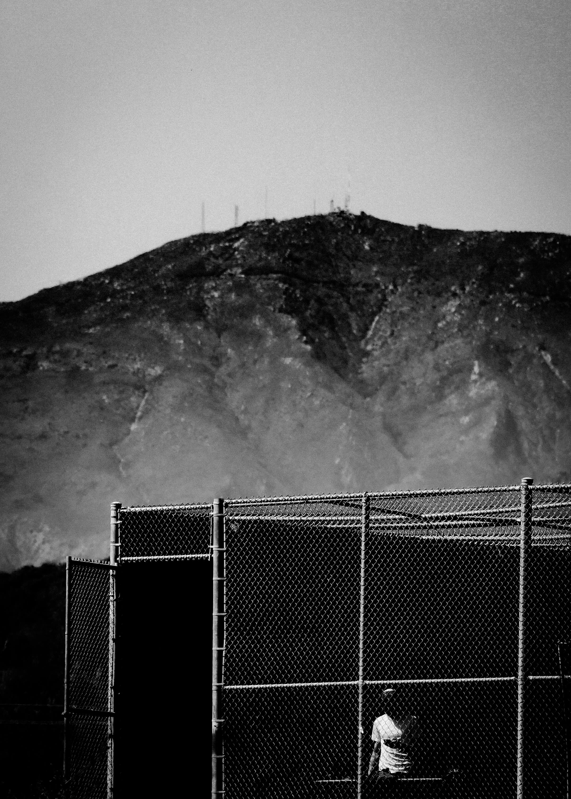 A black-and-white photograph of a mountain overshadowing a tennis court with a person inside